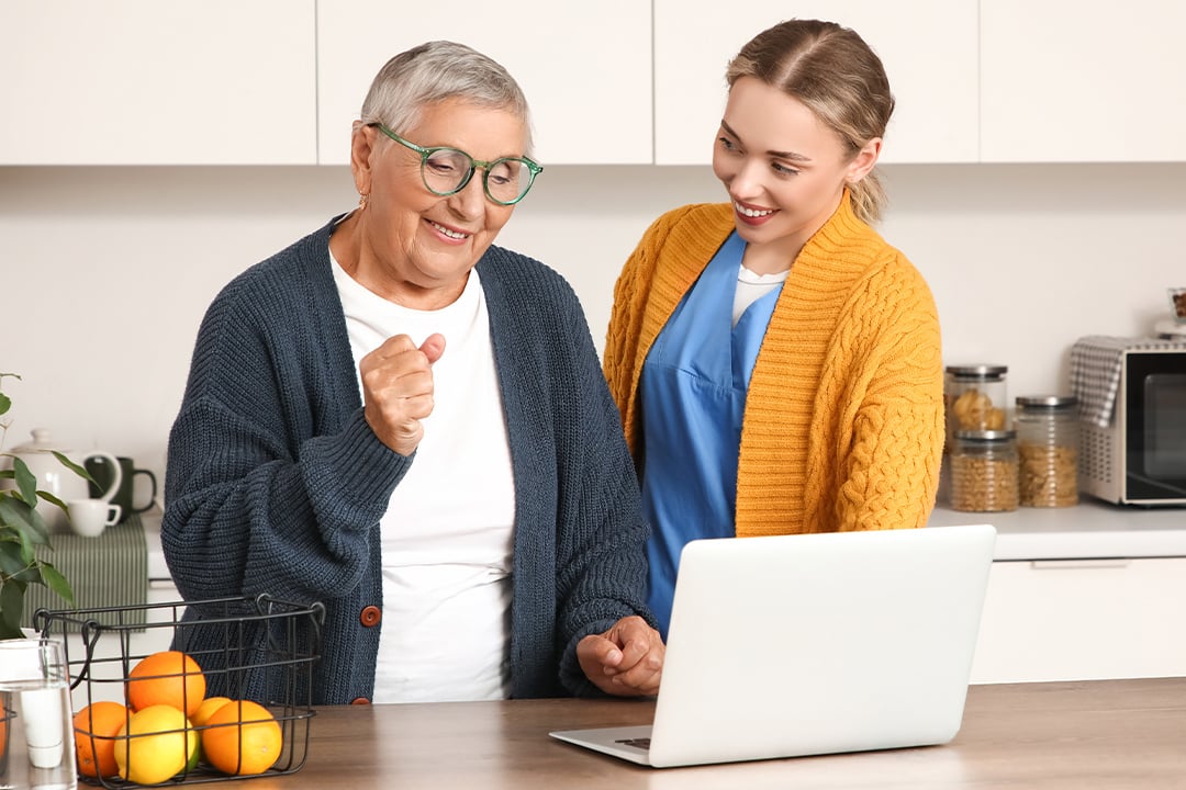 Older and younger adults smiling while looking at a laptop in a modern kitchen, symbolizing intergenerational tech engagement.
