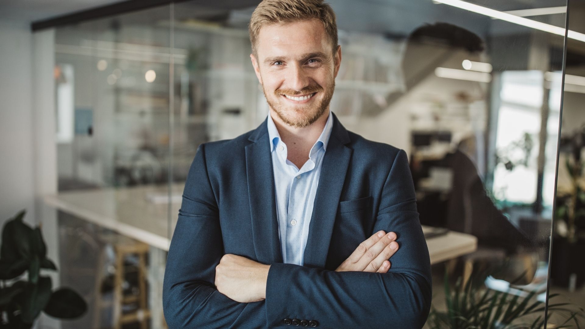 Smiling businessman in a blue suit standing with arms crossed in a modern glass-walled office.