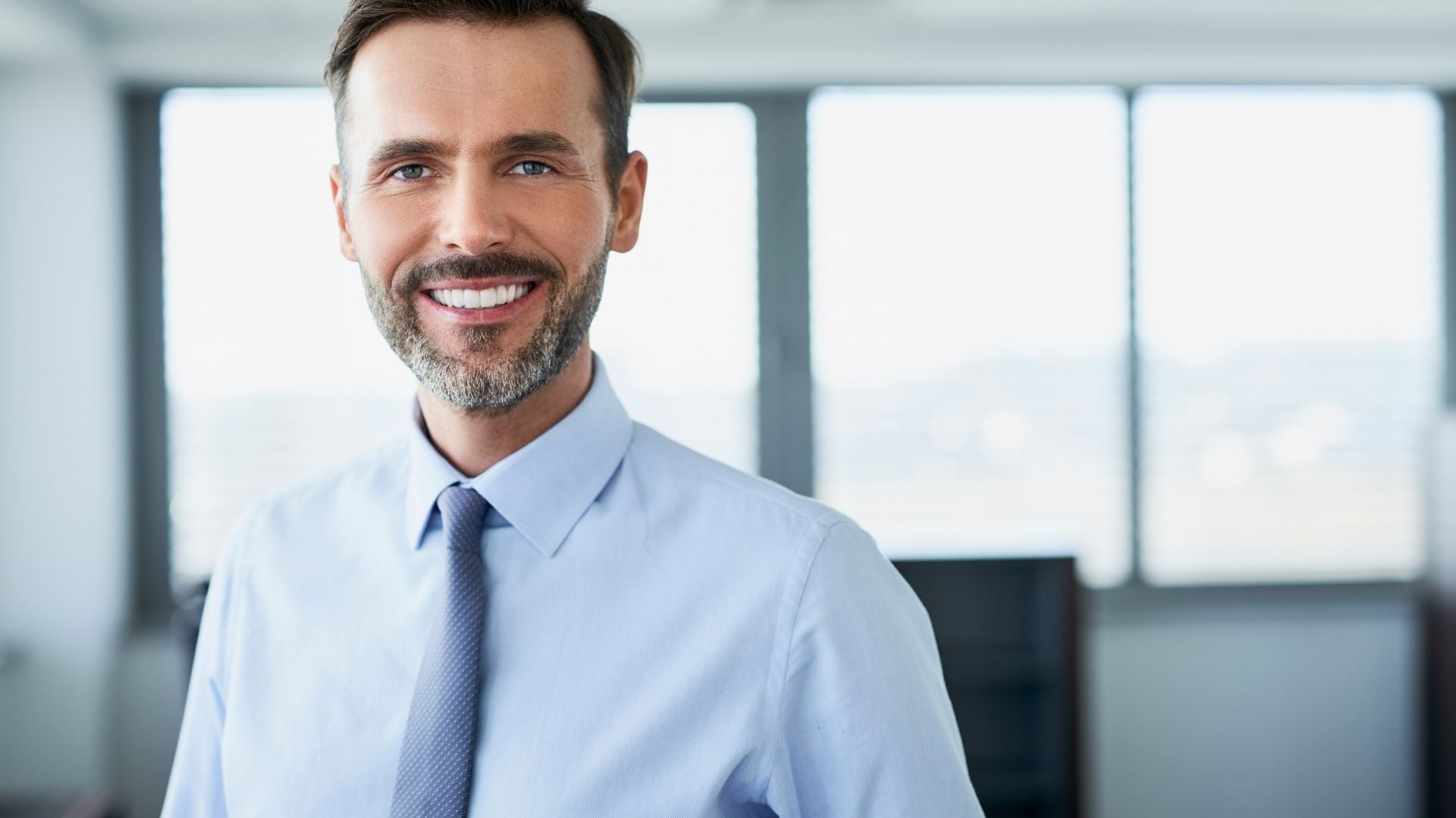 Smiling professional man in a dress shirt and tie standing in a bright office with large windows