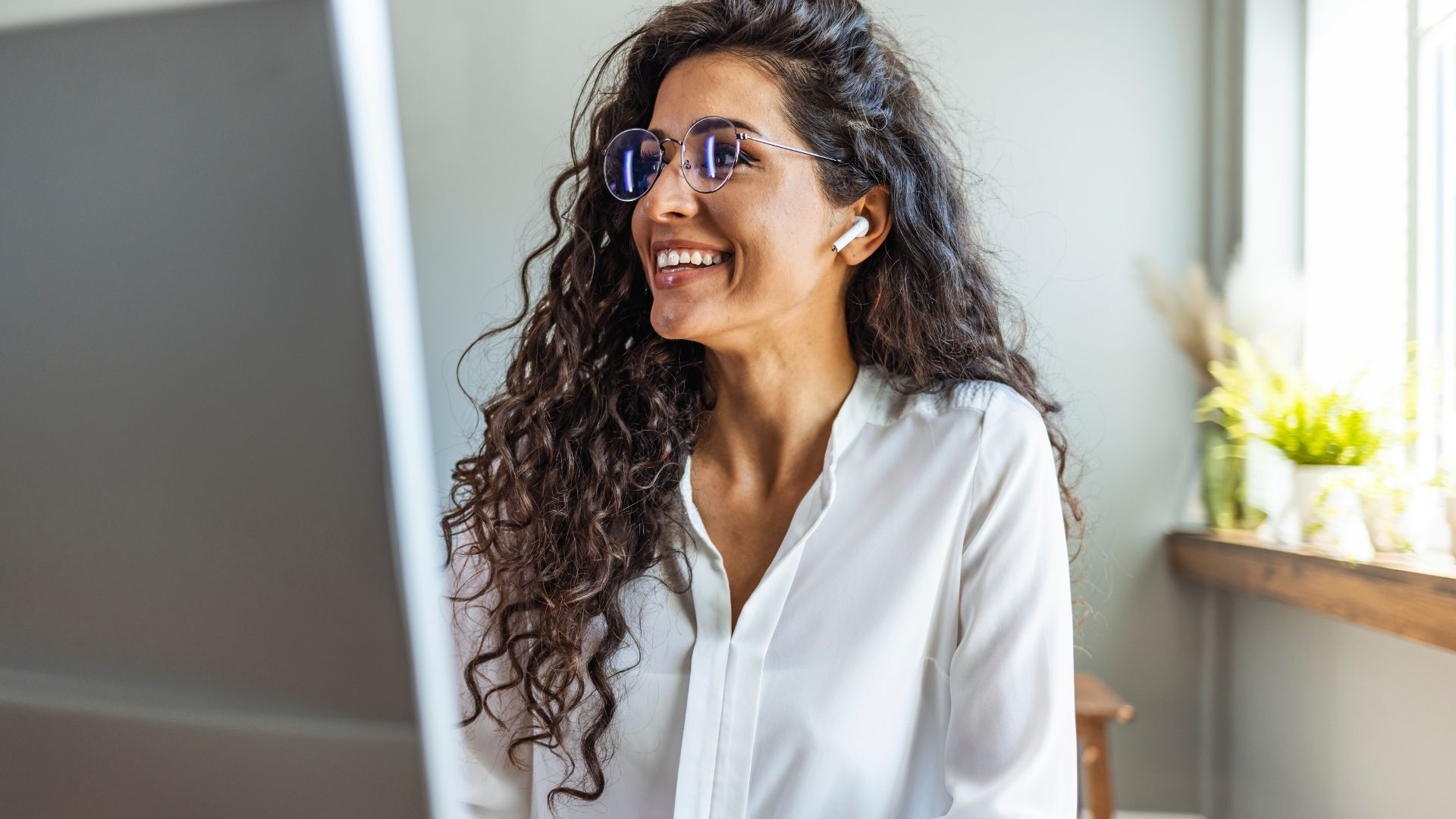 Smiling woman with curly hair and glasses wearing wireless earbuds, looking at a computer screen in a bright home office.