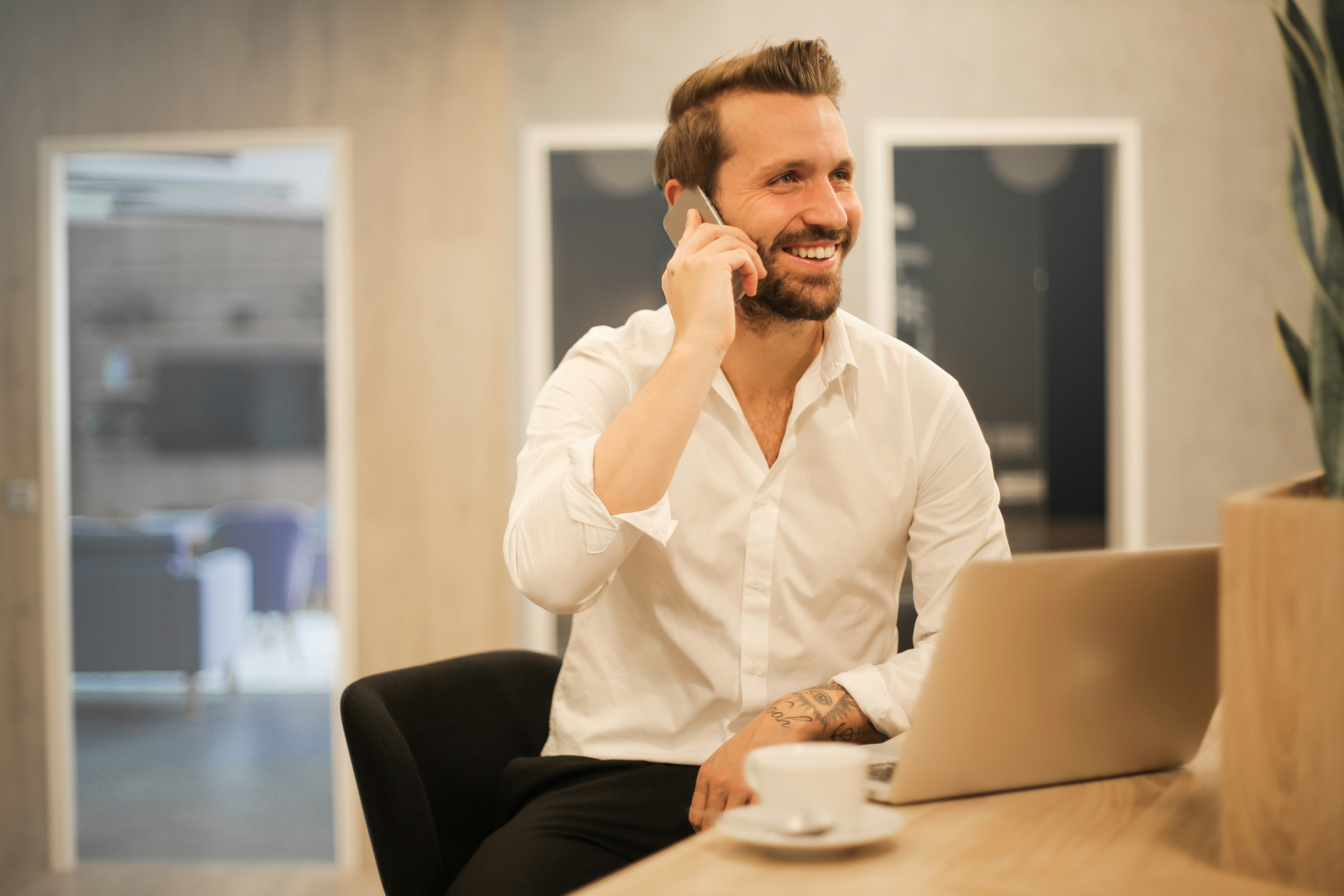 Smiling professional in a white shirt talking on the phone at a desk with a laptop and coffee cup in a relaxed, modern setting.